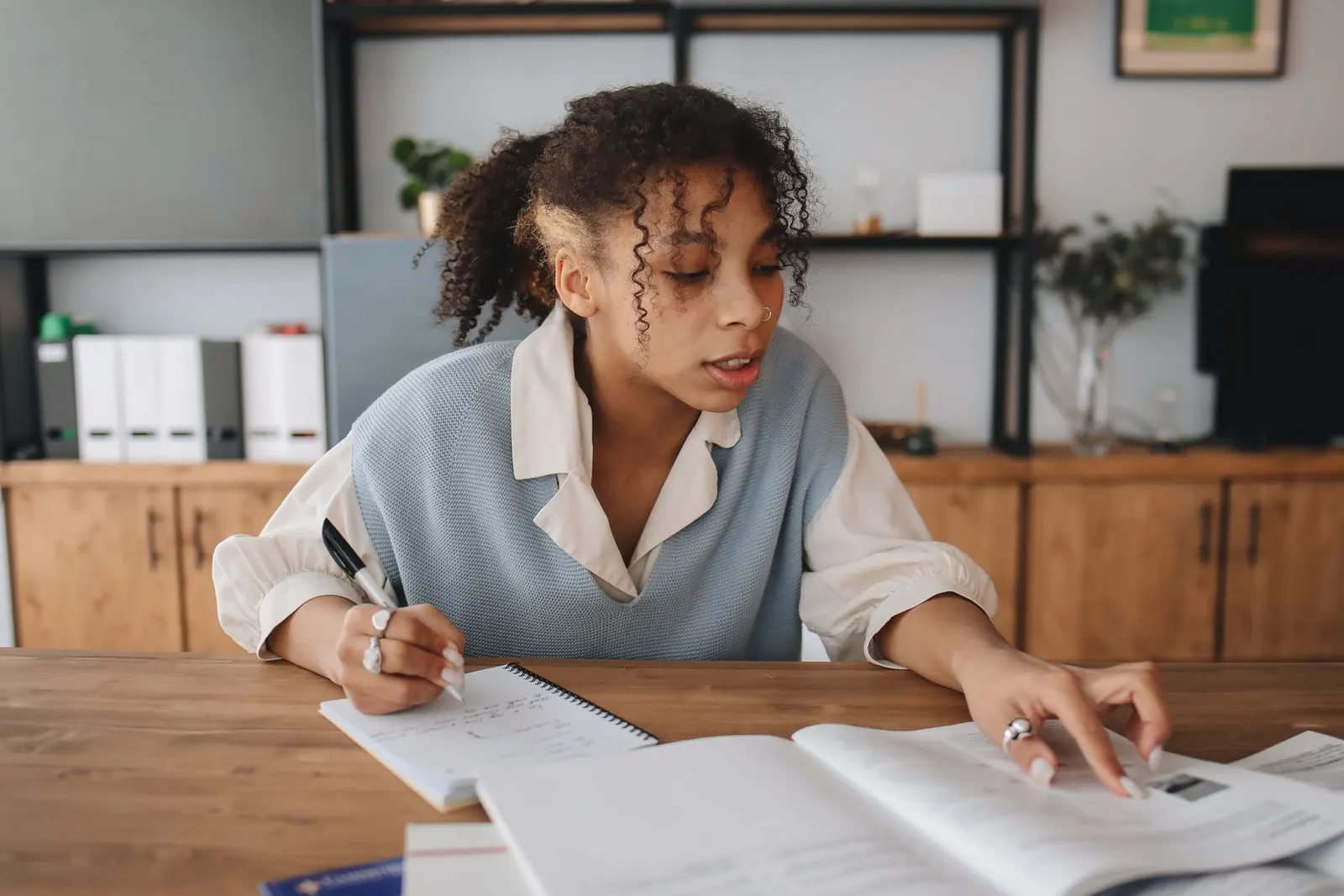A woman taking notes from a textbook at a wooden table while studying for a cybersecurity certification