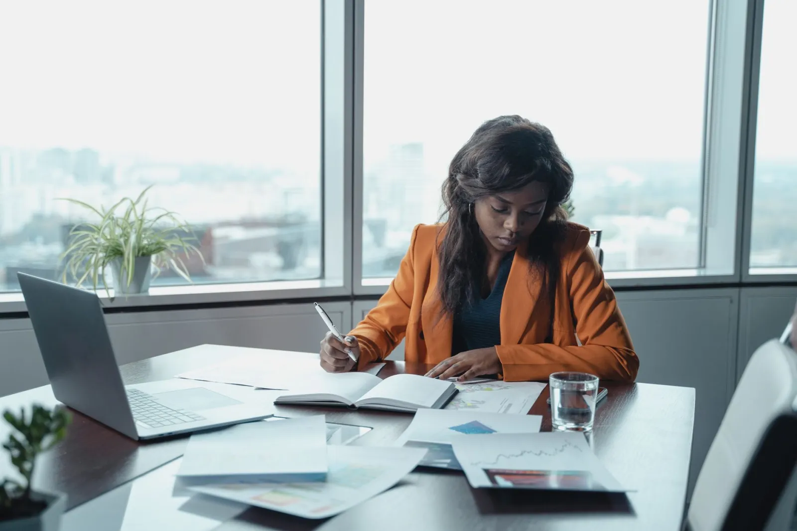 Businesswoman in an orange blazer taking notes over printed reports at a window desk