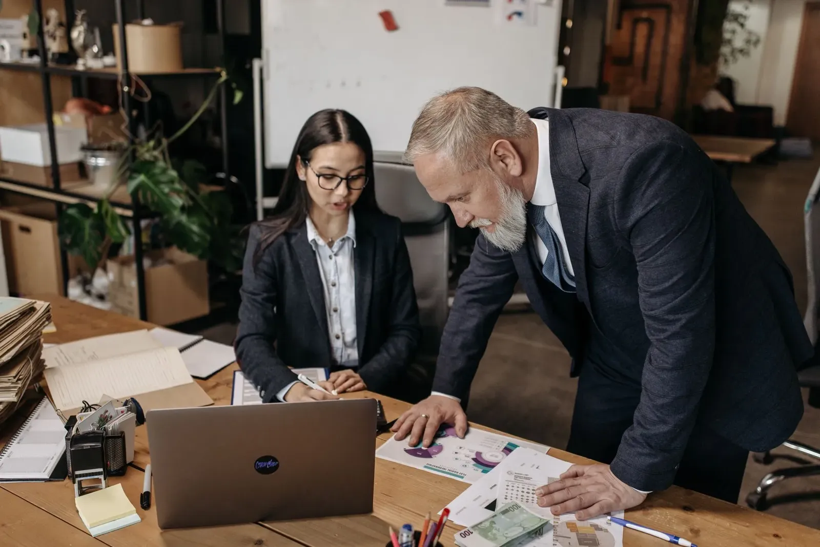 Senior manager in a suit reviewing printed charts with a colleague at a laptop in a busy office