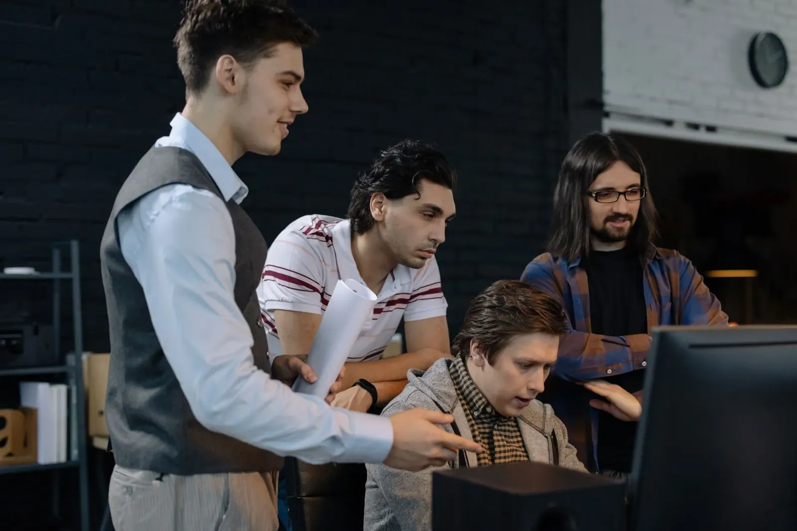 Team lead standing over a programmer's workstation as three coworkers review the screen together in a dark office