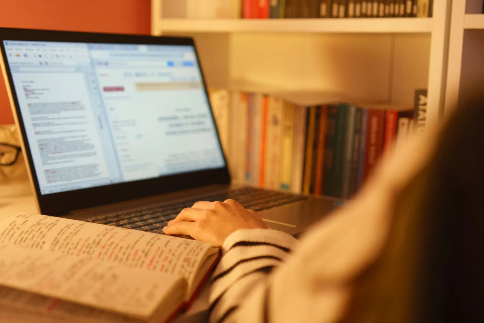 A student studying for an online cybersecurity degree at home with a laptop and open notes beside a bookshelf