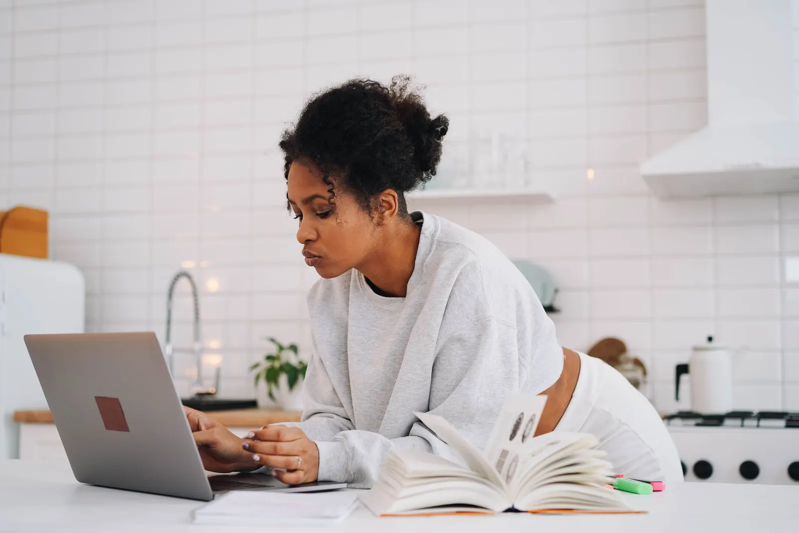 A woman researching online cybersecurity programs on a laptop with an open textbook in her kitchen