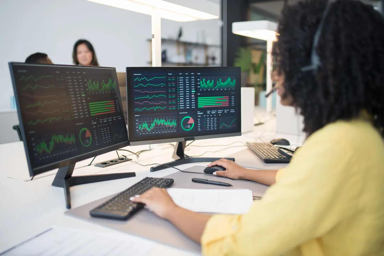 A woman at a desk reviewing data dashboards on two monitors, tied to cybersecurity salary data